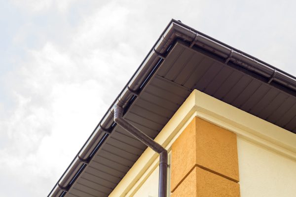 Close-up detail of cottage house corner with brown metal planks siding and roof with steel gutter rain system. Roofing, construction, drainage pipes installation and connection concept.